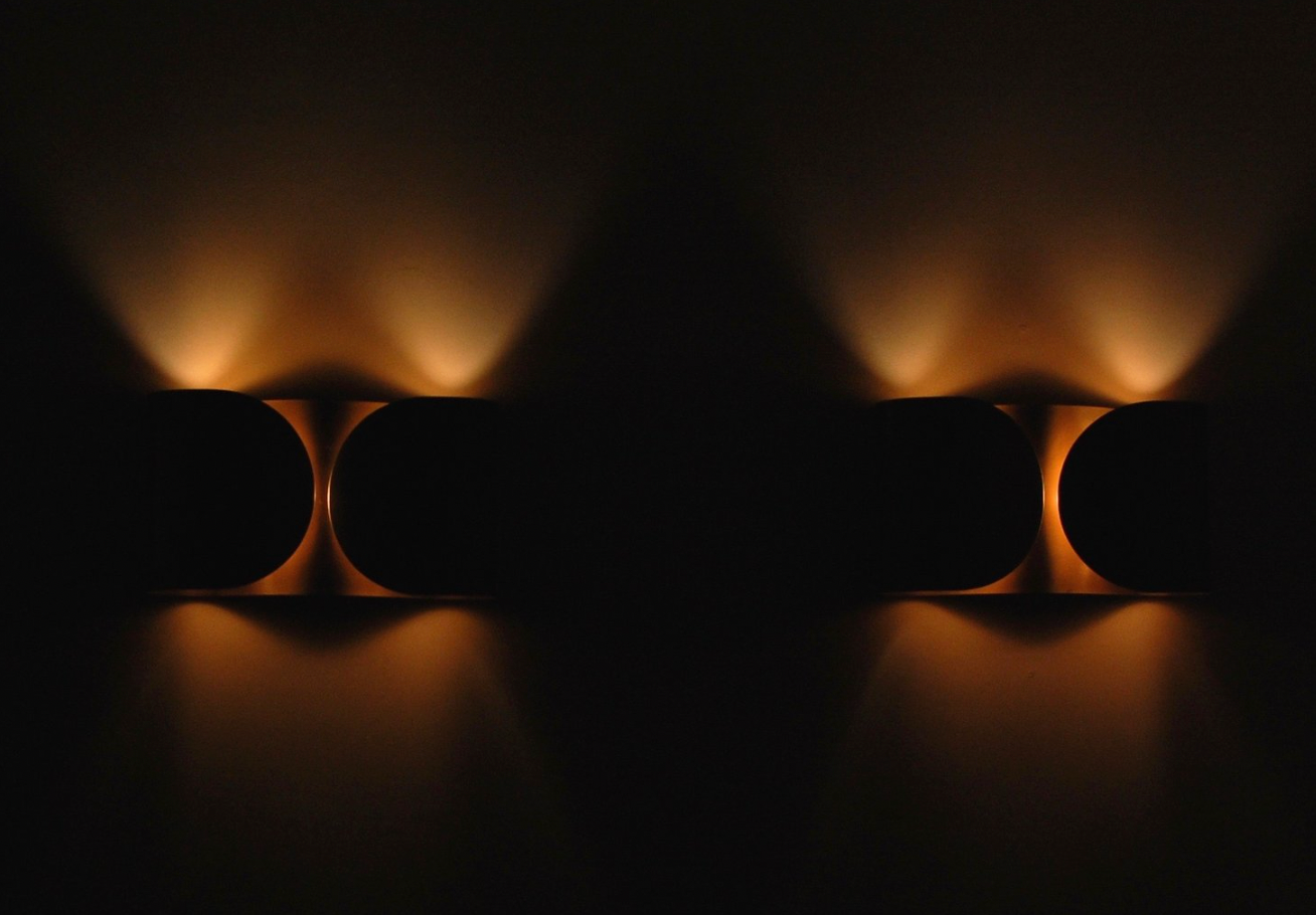 Postmodern brass wall sconces illuminated in a dark room, casting symmetrical up-and-down light in a sculptural Foglio-style pattern.