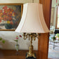 Vintage brass candelabra lamp with cream shade on wooden sideboard, styled with flowers and framed artwork in an elegant room.